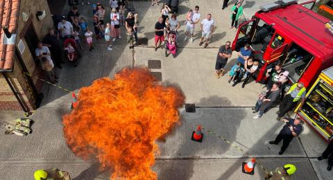 A drill yard at a fire station, full of people for an event, a fire engine and a large fire from a demonstration