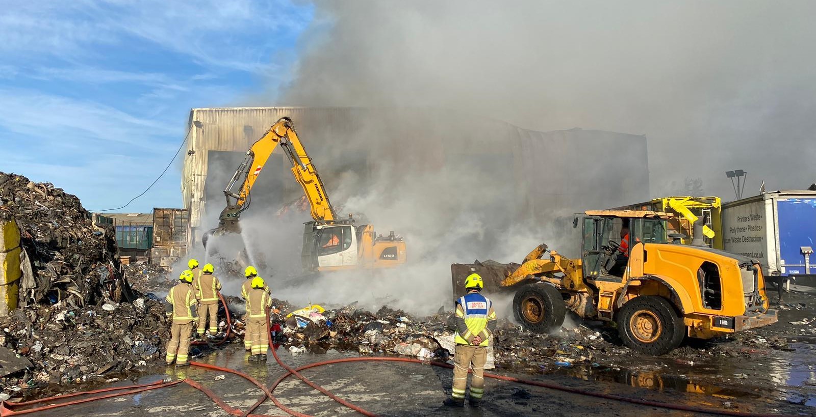 A digger breaking up a pile of scrap metal, with firefighters looking on