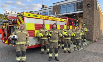 This is an image of eight firefighters, men and women, standing in their protective clothing in front of a fire engine. They are all smiling and holding their helmets in their arms.