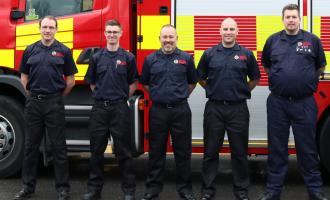 Five firefighters standing in front of a fire engine and smiling into the camera. 