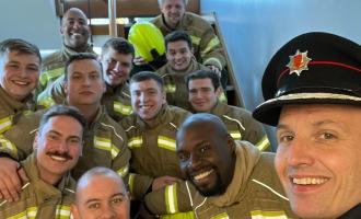A squad of eleven firefighters casually sitting on some steps and smiling at the camera, with Chief Fire Officer Rick Hylton at the front taking the selfie