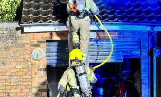 A firefighter climbing a ladder and a firefighter at the foot of the ladder, pitched against a garage