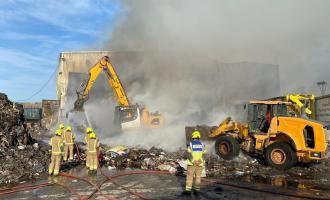 A digger breaking up a pile of scrap metal, with firefighters looking on