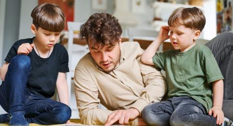 Dad and two young boys sat on a wooden floor, looking at a book together