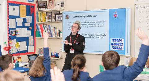 Education Officer standing by a projector in front of primary school children in blue uniform. One boy has raised his hand