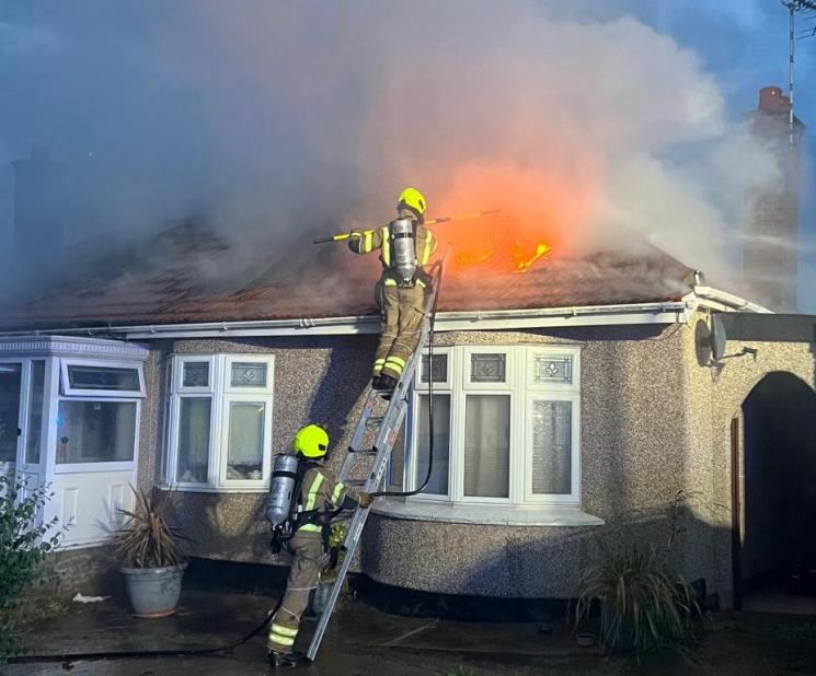 Smoke and flames coming out of the roof of a bungalow, with a firefighter on a ladder tackling the fire, while another firefighter holds the ladder