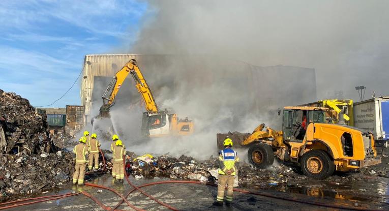 A digger breaking up a pile of scrap metal, with firefighters looking on